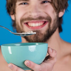 Happy man is having breakfast, eating porridge from bowl on blue background. Diet nutrition and healthy lifestyle, athlete's food, healthy food. The beginning of the day before work of the young guy